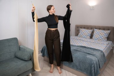 Genç bir kadın yatak odasında askıların üzerinde iki elbise tutuyor, hangisini giyeceğine karar vermeye çalışıyor, moda tercihleri kavramını gözler önüne seriyor ve hazırlanıyor.