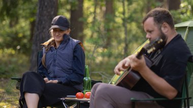 Sakallı adam ormanda kamp yaparken gülümseyen kadın için gitar çalıyor, çadır ve kamp ateşinin yanında katlanan sandalyelerde oturuyor, domatesler, salatalıklar ve katlanan masa üzerinde bir şişe birayla.