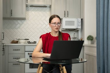 Gözlüklü ve kırmızı gömlekli genç bir kadın modern bir mutfakta cam bir masada oturuyor, bir dizüstü bilgisayarla işine odaklanıyor, muhtemelen uzaktan çalışıyor veya ev ekonomisini yönetiyor.