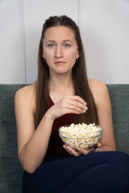 Genç bir kadın rahat bir koltukta oturmuş, oturma odasında film izlerken patlamış mısırın tadını çıkarıyor, rahat ve rahatlatıcı bir ortam yaratıyor. Dikey fotoğraf