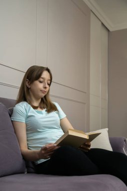 Evdeki rahat bir kanepede dinlenirken kitap tutan genç ve güzel bir kadın, sessiz mekanında huzur ve sükunetin tadını çıkarıyor. Dikey fotoğraf