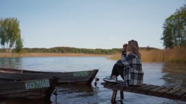 Ahşap iskelede oturan genç bir kadın güneşli huzurlu bir günün tadını çıkarıyor, sakin sulara bakıyor ve kıyıya demirlemiş eski teknelere bakıyor, güzel doğal manzara Belarus Braslav 'ı düşünüyor.