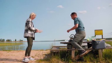 Genç mutlu çift göl kenarında piknik masası kuruyor, serin bir çantadan yiyecek indiriyor ve doğada güneşli bir günde taşınabilir gaz ocağında sıcak bir içecek hazırlıyor.