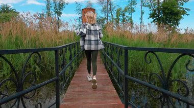 Güzel bir yaz parkında, uzun yeşil sazlıklar ve ağaçlarla çevrili bir göletin üzerindeki ahşap köprüde koşan, ekose gömlekli kaygısız sarışın bir kadının arka görüntüsü.