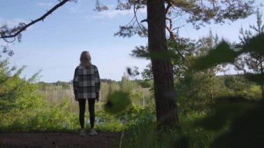 Arkası kameraya dönük genç bir kadın manzaralı bir manzarada hareketsiz duruyor, yemyeşil bir ormanın güzel manzarasına ve güneşli bir yaz gününde sakin bir göle hayran.