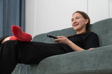 Kırmızı çorap giyen genç bir kadın oturma odasında rahat bir koltukta oturuyor, uzaktan kumandayı tutuyor ve televizyonda komedi programı izlerken gülüyor.