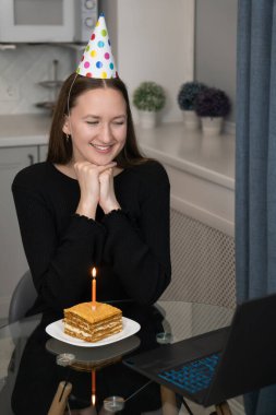 Parti şapkası takan genç bir kadın doğum günü pastasındaki mumu üflemeden önce bilgisayarını kullanarak arkadaşları ve ailesiyle internette kutlama yapıyor. Dikey fotoğraf