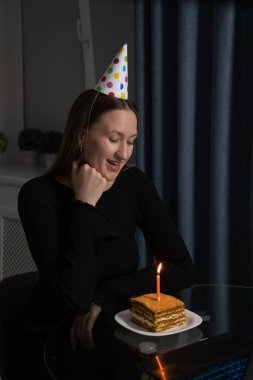 Genç bir kadın doğum gününü internette kutluyor, şenlikli bir parti şapkası takıyor ve üzerinde mumla küçük bir pastayla mutfağında oturuyor, neşeli bir anın tadını çıkarıyor. Dikey fotoğraf