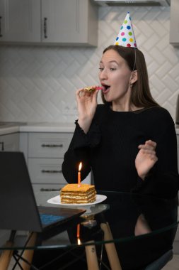 Genç bir kadın internetten doğum gününü kutluyor, parti şapkası takıyor ve parti kornası çalıyor, mutfakta dizüstü bilgisayarla pasta yerken bir dilim mumun keyfini çıkarıyor.
