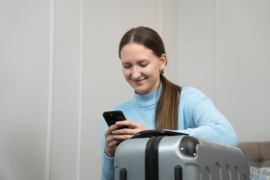 Eve gitmek için bekleyen genç bir kadın, akıllı telefonunu kullanarak seyahat ayarlamaları yapıyor. Bu sırada pasaportu ve bavulu yakınlarda oturuyor ve yaklaşan yolculuk için heyecan saçıyor.