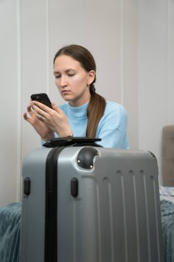 Genç bir kadın bavulunun yanında oturuyor, akıllı telefonunu seyahat hazırlıkları için kullanıyor, yaklaşan seyahatini planlıyor ve online rezervasyonları ayarlıyor. Dikey fotoğraf