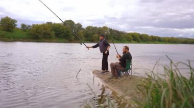 Birkaç erkek ve kadın kumlu nehir kıyısında dinlenmenin keyfini çıkarıyorlar: biri ayakta ve atış halatı, diğeri olta kamışı ile katlanır bir sandalyede oturuyor.