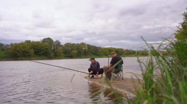 Birkaç erkek ve kadın kumlu nehir kıyısında dinlenmenin keyfini çıkarıyorlar: biri ayakta ve atış halatı, diğeri olta kamışı ile katlanır bir sandalyede oturuyor.