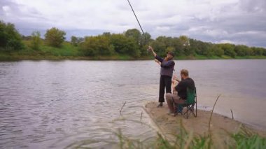 Birkaç erkek ve kadın kumlu nehir kıyısında dinlenmenin keyfini çıkarıyorlar: biri ayakta ve atış halatı, diğeri olta kamışı ile katlanır bir sandalyede oturuyor.
