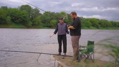 Nehir kıyısında bir kadın balık oltası atarken bir adam sandalyede onun yanında oturuyor ve güzel, doğal bir ortamda birlikte balık tutmanın keyfini çıkarıyor.