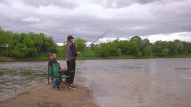 Genç bir kadın nehir kıyısından gelen bir oltayla balık tutuyor ve erkek arkadaşı da bulutlu bir günde kamp sandalyesinde oturup dinleniyor.