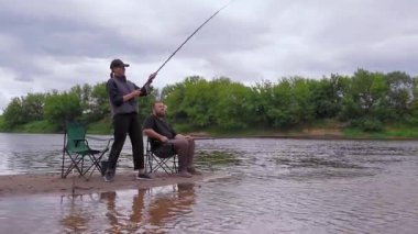 Nehir kıyısında bir kadın balık oltası atarken bir adam sandalyede onun yanında oturuyor ve güzel, doğal bir ortamda birlikte balık tutmanın keyfini çıkarıyor.