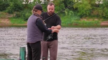Orta yaşlı sakallı bir adam sabırla genç bir kadına balık oltasının üzerinde makarayı nasıl kullanacağını öğretiyor. Ortak hobileri için oltayı çözmesine yardım ediyor.