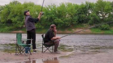 Genç çift, birlikte sakin bir gün geçirmenin keyfini çıkarırken, kadın ayakta durup oltasını nehre atarken, adam düşünceli bir şekilde bir kamp sandalyesinde oturuyor.
