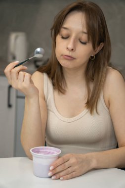 Genç bir kadın mutfak masasında oturuyor, kahvaltıda yoğurdun tadını çıkarıyor, sabah rutininin ayrılmaz bir parçası olarak sağlıklı bir yaşam tarzını benimsiyor ve genel refahı destekliyor.