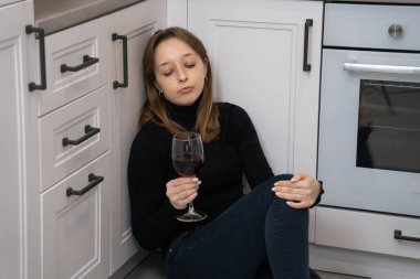 Genç bir kadın mutfak zemininde oturmuş şarap yudumluyor, yalnızlık ve üzüntü ile boğuşuyor, duygusal karmaşayla boğuşuyor.