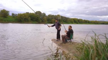 Birkaç erkek ve kadın kumlu nehir kıyısında dinlenmenin keyfini çıkarıyorlar: biri ayakta ve atış halatı, diğeri olta kamışı ile katlanır bir sandalyede oturuyor.