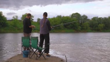 Birkaç erkek ve kadın kumlu nehir kıyısında dinlenmenin keyfini çıkarıyorlar: biri ayakta ve atış halatı, diğeri olta kamışı ile katlanır bir sandalyede oturuyor.