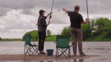 Birkaç erkek ve kadın kumlu nehir kıyısında dinlenmenin keyfini çıkarıyorlar: biri ayakta ve atış halatı, diğeri olta kamışı ile katlanır bir sandalyede oturuyor.