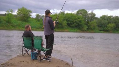 Birkaç erkek ve kadın kumlu nehir kıyısında dinlenmenin keyfini çıkarıyorlar: biri ayakta ve atış halatı, diğeri olta kamışı ile katlanır bir sandalyede oturuyor.