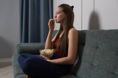 Genç bir kadın oturma odasındaki rahat bir koltukta oturmuş, bir kase patlamış mısırla film gecesinin tadını çıkarıyor, ekrandaki eğlenceye tamamen dalmış.