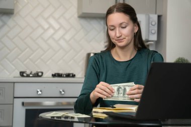 Genç bir kadın ev ekonomisini yönetiyor, kalan dolarları sayıyor, dizüstü bilgisayarla mutfak masasında oturuyor, maddi sorunlar yüzünden stresli hissediyor ve faturaları ödemekte zorlanıyor.