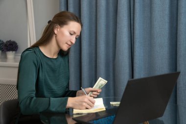 Konsantre kadın serbest çalışan titizlikle finansını yönetiyor, ev ofisinde çalışıyor, gelir ve giderleri hesaplarken bir dizüstü bilgisayar ve defter kullanıyor.