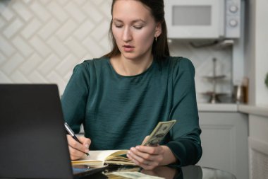 Bize dolar banknotları sayarak ev bütçesini yöneten, mutfaktaki cam masada oturan, dizüstü bilgisayarda çalışan ve not defterine notlar yazan genç bir kadın.