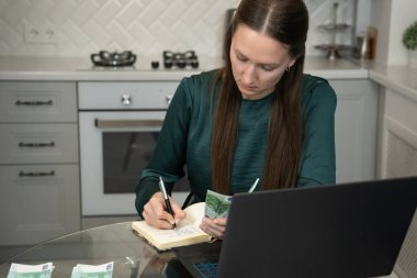 Kadın dikkatli bir şekilde ev ekonomisini yönetiyor. Titizlikle bir deftere harcamalarını yazıyor. Avro banknotlarını tutuyor ve mutfaktaki dizüstü bilgisayarını kullanıyor.