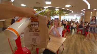 Minsk, Belarus - 13 Temmuz 2024: POV, bir alışveriş merkezinin gürültülü yemek katındaki masada oturan mutlu sarışına yemek getiren adamı fast food ile kutlarken çekti.