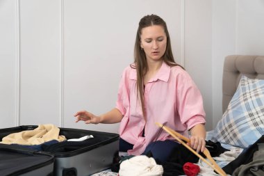 Yolculuk için giysilerini toplayan genç bir kadın, evdeki yatakta oturuyor, giysileri bir bavula yerleştiriyor, seyahate ya da tatile hazırlanıyor.