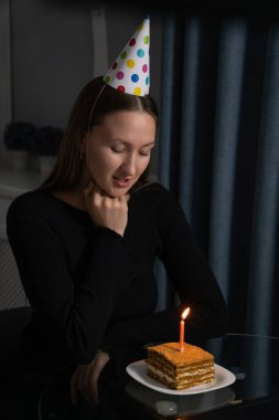 Renkli bir parti şapkası olan genç bir kadın, kapalı gözlerle ve kapalı ellerle bir dilek diliyor, sıcak bir masada doğum gününü pasta ve yanan bir mumla kutluyor. Dikey fotoğraf