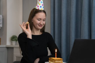 Parti şapkası takan gülümseyen genç bir kadın, doğum gününü mutfağında, önünde bir doğum günü pastası ve yanan bir mumla kutlarken parti düdüğü çalıyor.