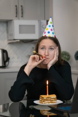 Üzgün yalnız kadın parti şapkası takıyor ve doğum gününü internetten kutluyor dizüstü bilgisayarla, mutfak masasında doğum günü pastası ve mum yakıyor. Dikey fotoğraf