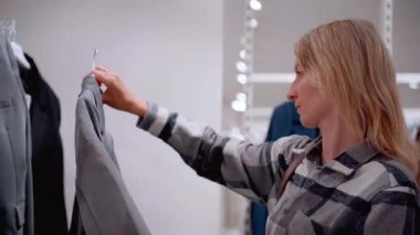 Genç sarışın kadın modern bir giyim mağazasından şık bir ceket seçiyor. Mükemmel bir iş elbisesi ya da hediye aramak için yeni bir koleksiyona göz atıyor.