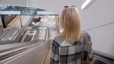Sarı saçlı, ekose gömlek giyen ve omuz çantası taşıyan, modern ve boş bir metro istasyonundaki yürüyen merdivenden tren istasyonuna inen genç bir kadının arka görüntüsü.