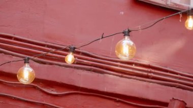 Şehir manzarasını aydınlatan bir çelenk üzerindeki sıcak, klasik ampuller, eski pencereli tarihi kırmızı bir binanın önünde nazikçe sallanarak, samimi ve şenlikli bir atmosferik zemin oluşturur.