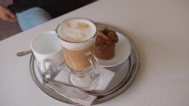 Şeffaf cam bardakta süt köpüklü enfes sıcak kapuçino. Küçük bir çikolatalı pasta ve sıcak bir kahve dükkanında bir bardak su ile servis edilir.