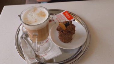 Bir bardak kapuçino ve kafedeki parlak metal tepside tatlı bir çikolatalı pasta ile nefis bir kahvaltı, rahatlatıcı bir mola için mükemmel bir tatlı ve sıcak aromatik bir içecek.