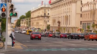 Minsk, Belarus - 13 Temmuz 2024: İstikrarlı araba trafiğiyle dolu şehir caddesi, yaya geçidinde bekleyen yaşlı bir kadın, caddeyi kaplayan tarihi binalar