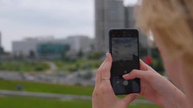 Arka görüş sarışını cep telefonu kullanarak modern şehir binalarının fotoğraflarını çekiyor, şehir mimarisini yakalıyor ve manzaralı perspektiften sosyal medyada anı paylaşıyor.