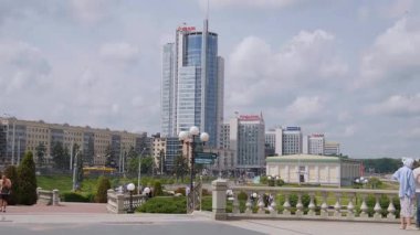 Minsk, Belarus - 13 Temmuz 2024: Bulutlu gökyüzüne karşı modern cam gökdelenin yer aldığı Minsk şehrinin manzaralı manzarası, yayalar ön planda kayalıklarda geziniyor.