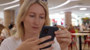 Sarışın, akıllı telefonunun beslemesini dikkatle izleyen ve kaydıran, kalabalık bir yemek salonunda oturan, dijital dünyasına dalmış bulanık insanların ve ışıkların arka planına bakan