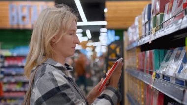 Düşünceli bir kadın marketteki raftan bir çikolata çubuğu seçer, şeker reyonunda karar vermeden önce etiketi ve malzemeleri dikkatle okur.