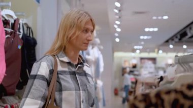 Ekose gömlekli genç sarışın kadın moda mağazasından elbise seçiyor alışverişin tadını çıkarıyor ve kendisi için mükemmel kıyafeti arıyor.
